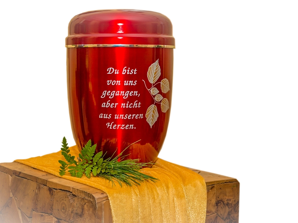 Eine glänzende, rote Urne mit der Inschrift "Du bist von uns gegangen, aber nicht aus unseren Herzen" und filigranen Blattmotiven, platziert auf einem Holzsokel mit goldenem Tuch und Grünschmuck. Bestattungsfotografie von Michael Schalansky.